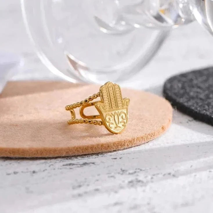 Gold Hamsa hand ring on a wooden coaster with a glass in the background