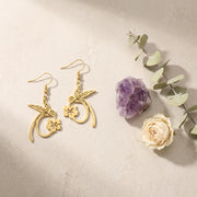 Gold earrings with floral designs on a beige surface with a purple crystal and white rose.