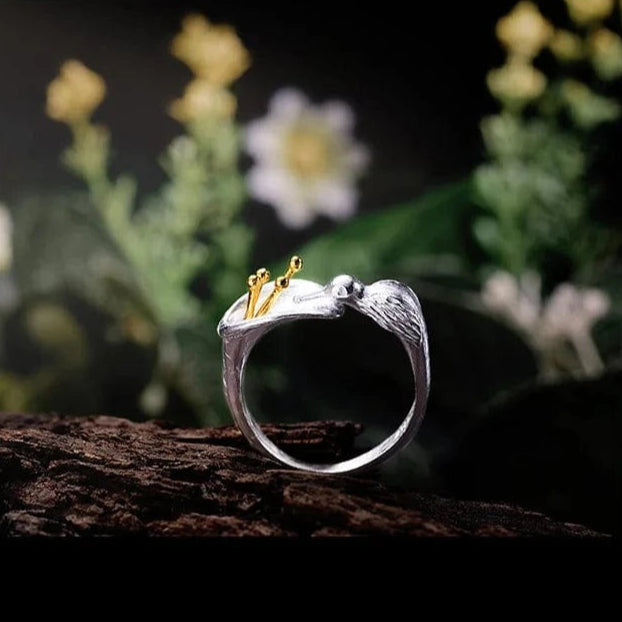 Silver ring with a hummingbird design on a wooden surface with flowers in the background