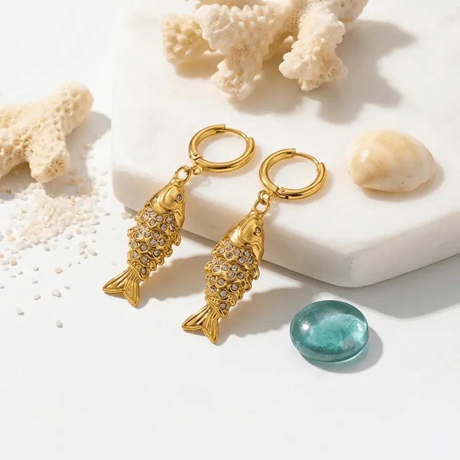 Gold fish-shaped earrings with gemstones on a white surface with coral and shells.