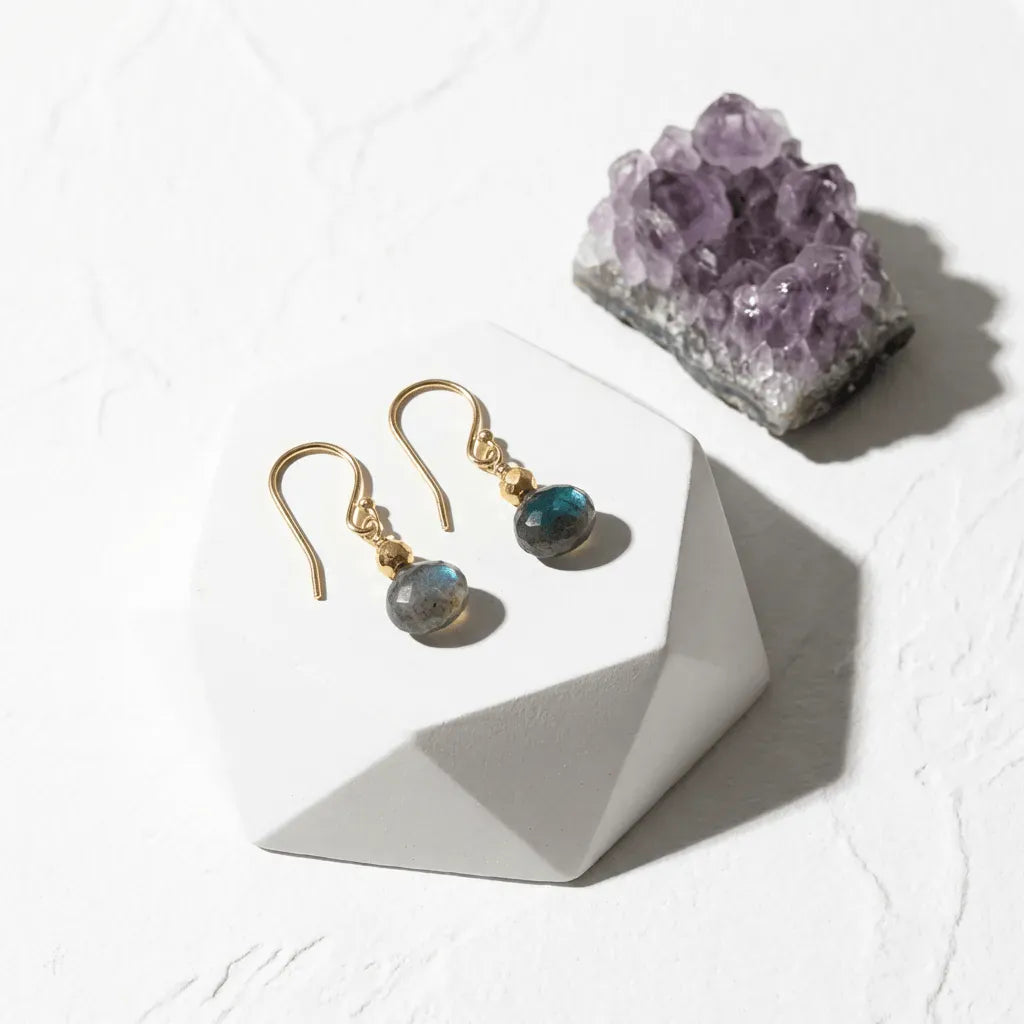 Gold earrings with gemstones on a white surface next to an amethyst crystal.
