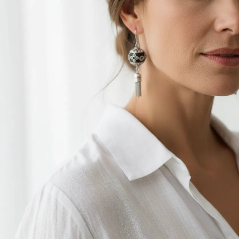 A close-up of a woman wearing a dangling earring with a silver-toned ball featuring black floral detailing and a silver tassel