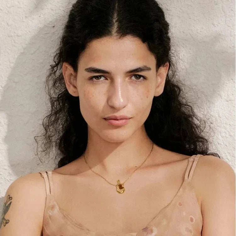 Woman with long dark hair and tattoos on her arm, wearing a sheer top against a textured white background.