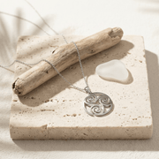 Silver necklace with a triskele pendant on a stone surface with a piece of driftwood and a white stone.