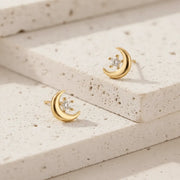 Gold moon-shaped earrings with embedded diamonds on a textured beige surface