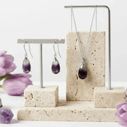 Jewelry display with purple teardrop earrings and necklace on a stone stand with flowers.
