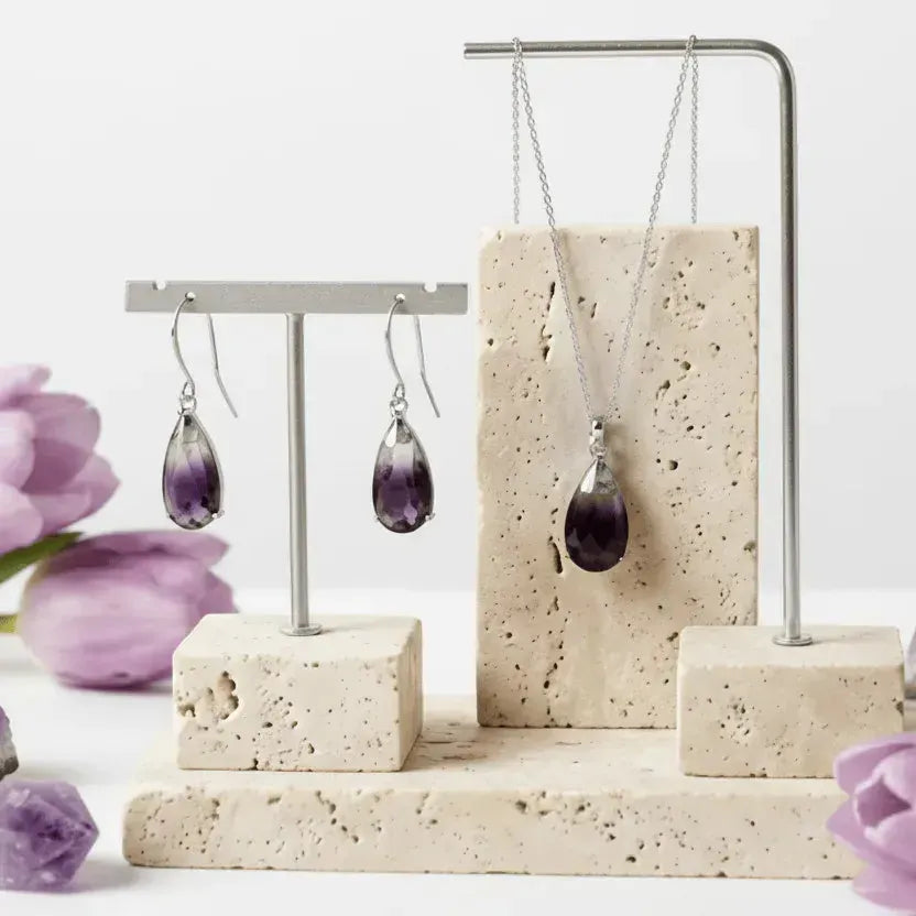 Jewelry display with purple teardrop earrings and necklace on a stone stand with flowers.
