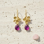 Gold flower earrings with purple teardrop stones on a textured beige surface.