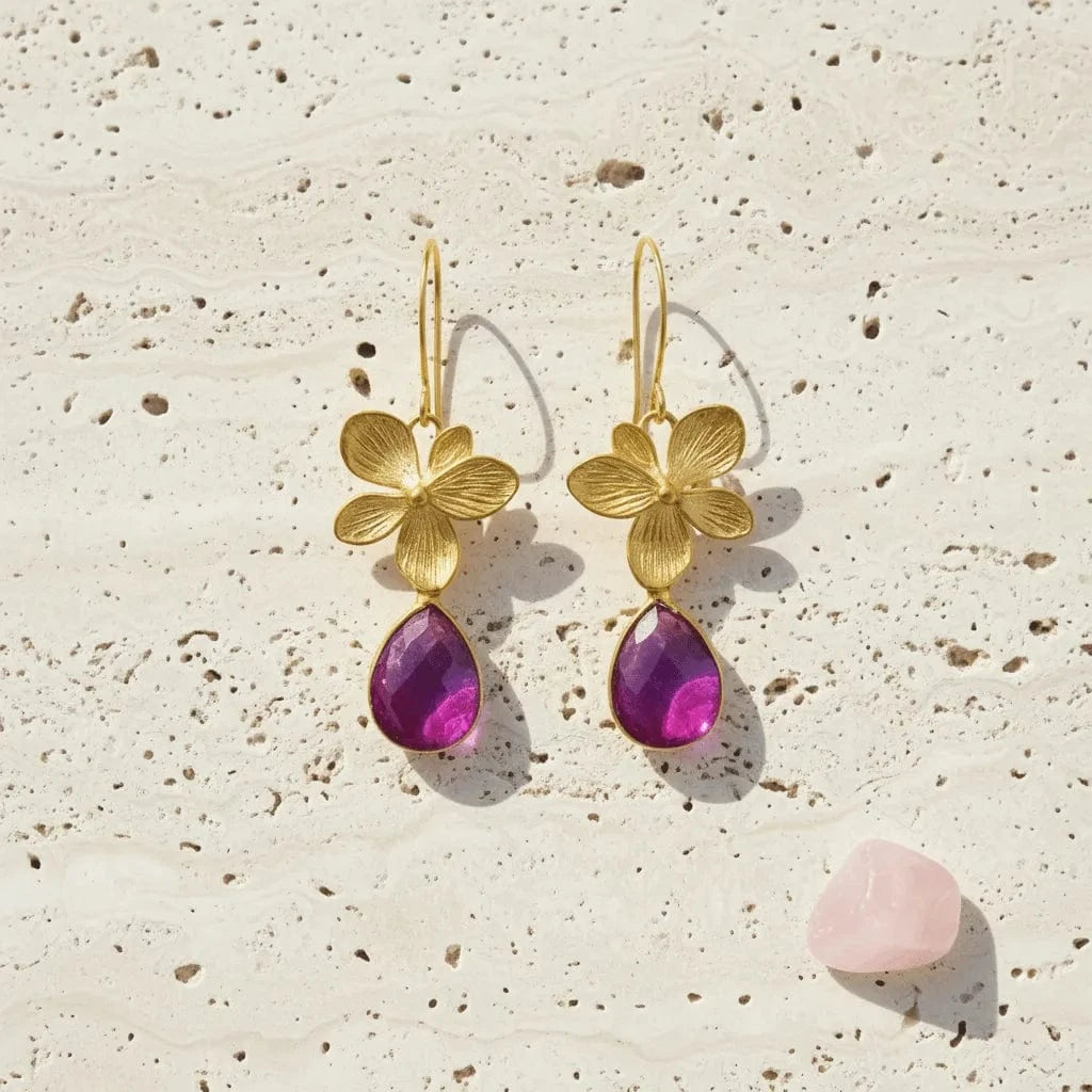 Gold flower earrings with purple teardrop stones on a textured beige surface.