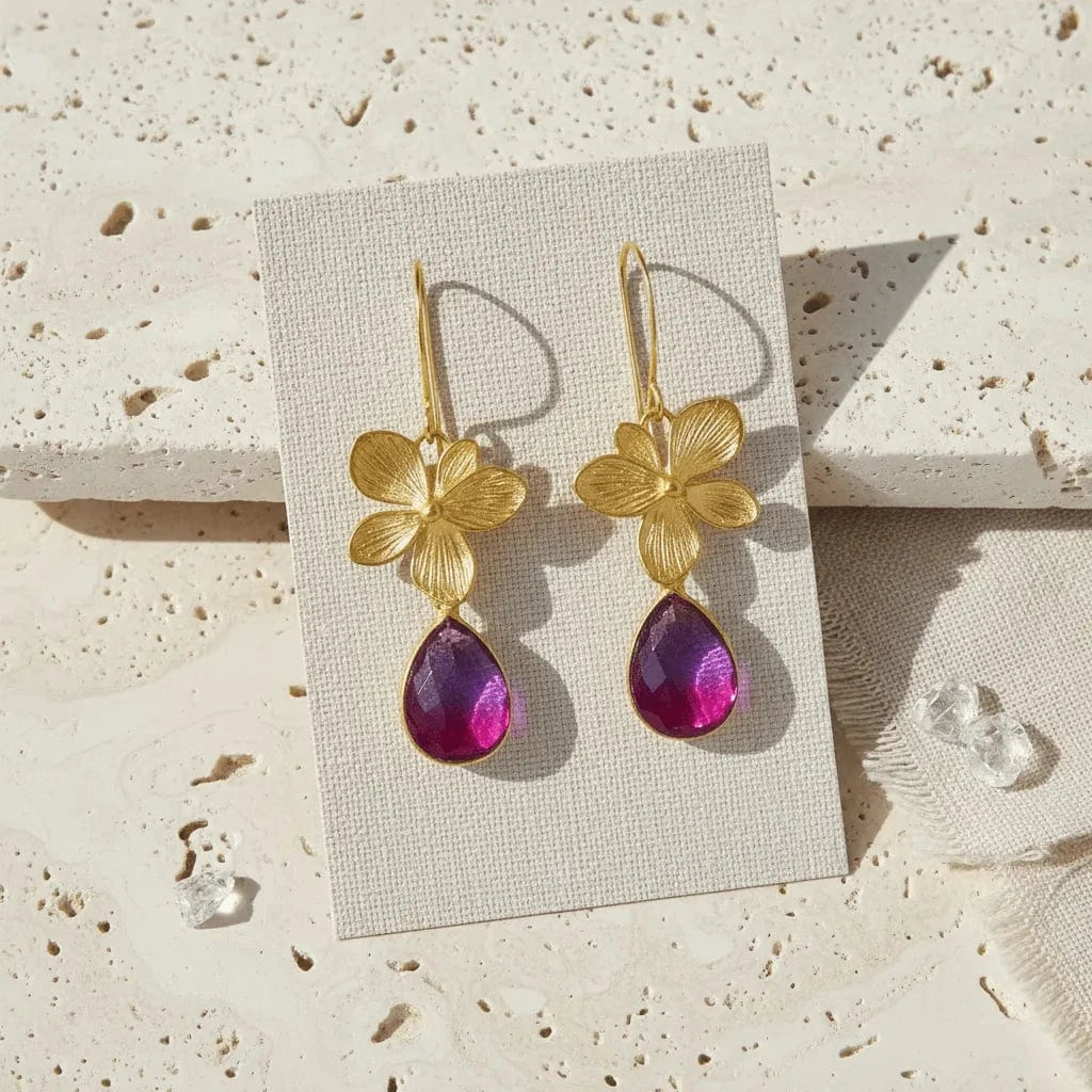 Gold floral earrings with purple teardrop stones on a beige card against a textured stone background