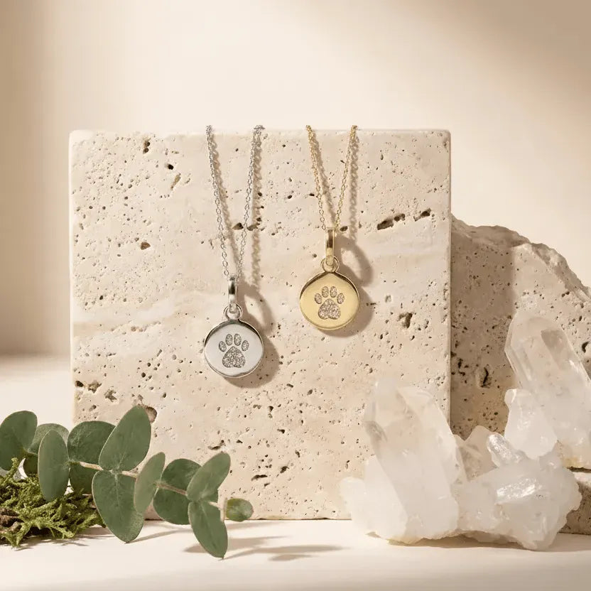 Two paw print necklaces, one silver and one gold, on a stone surface with crystals and eucalyptus leaves.
