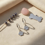 Pair of silver paw print earrings on a beige surface with lavender and a pink stone.