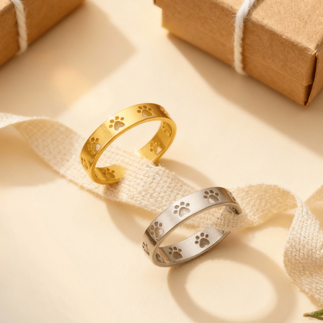 Gold and silver paw print rings on a beige fabric background with a brown gift box.