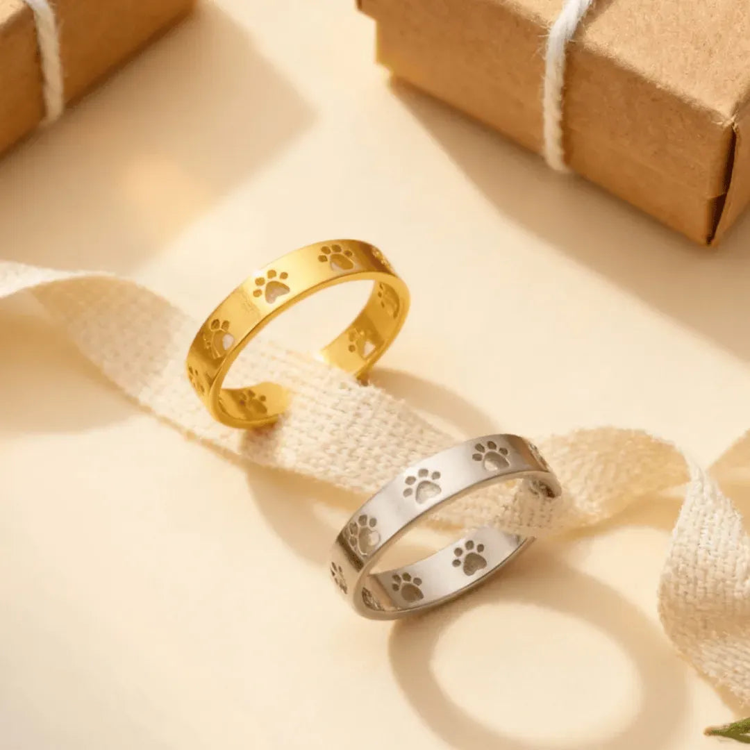 Gold and silver paw print rings on a beige fabric background with a brown gift box.
