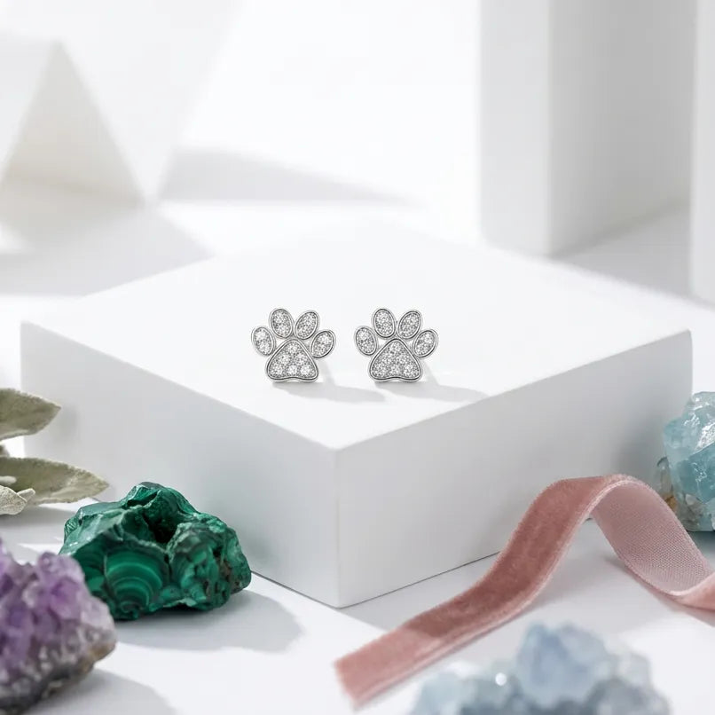 Silver paw-shaped earrings on a white surface with crystals and a pink ribbon.