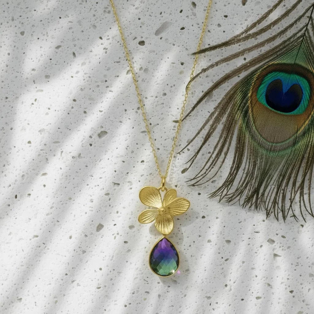 Gold necklace with a floral pendant and teardrop-shaped gemstone next to a peacock feather on a textured surface.