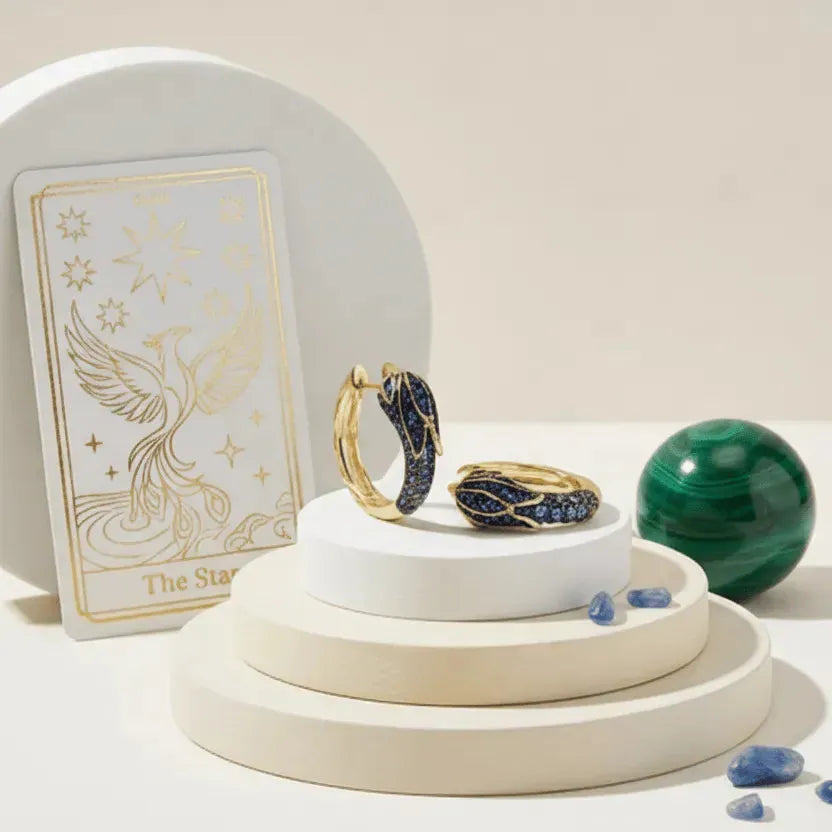 Gold hoop earrings with blue stones on a white stand, tarot card, and green crystal ball in the background.