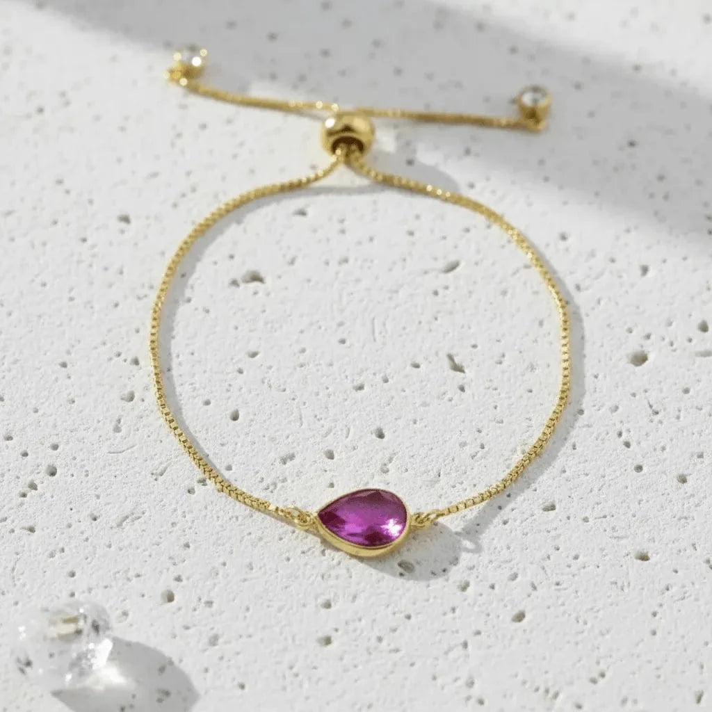 Gold bracelet with a purple teardrop-shaped gemstone on a textured white surface