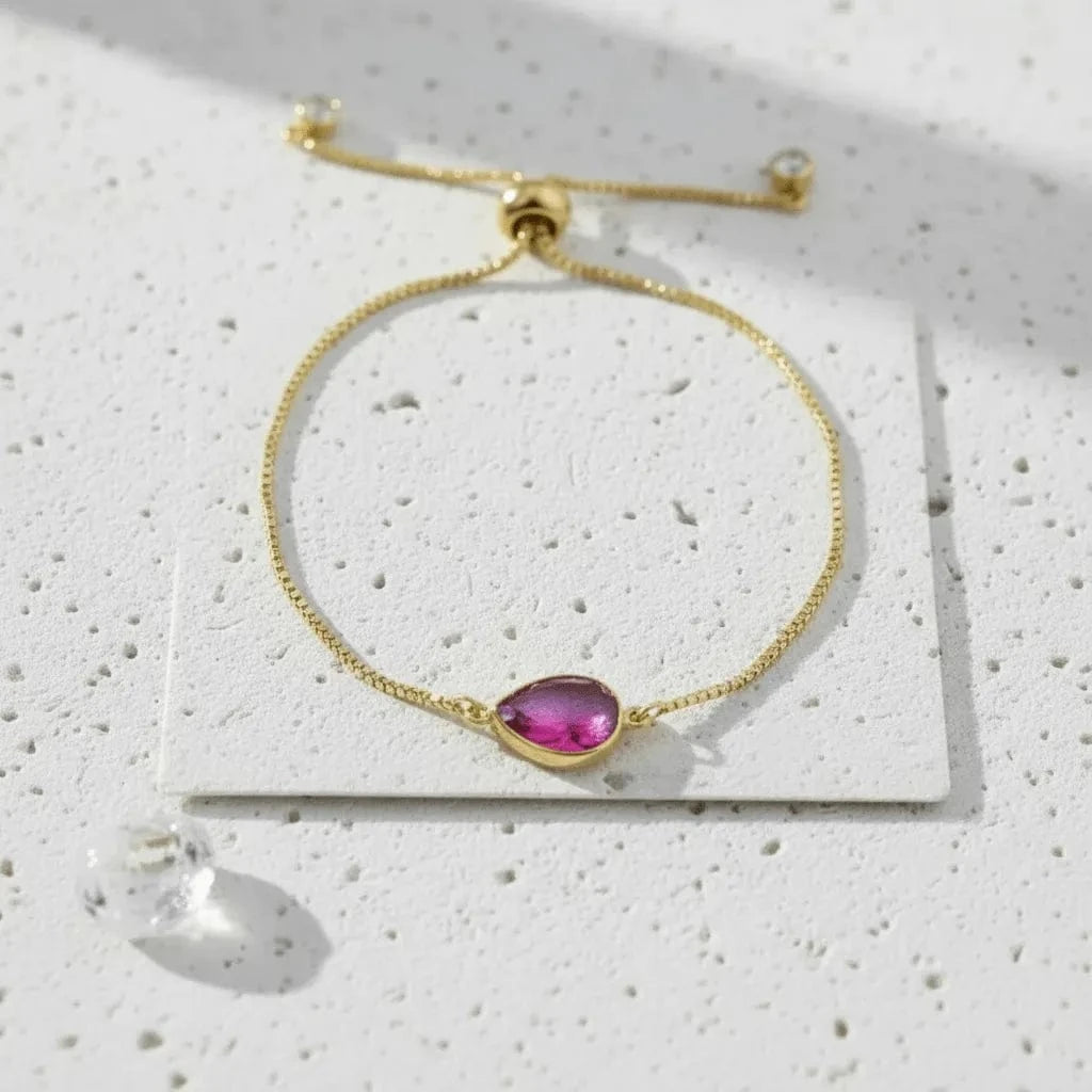 Gold bracelet with a purple teardrop-shaped gemstone on a textured white surface