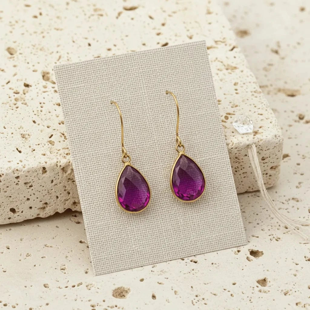 Purple teardrop earrings with gold hooks on a beige card against a textured beige background