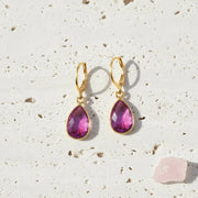 Gold earrings with pink teardrop-shaped gemstones on a textured beige surface.