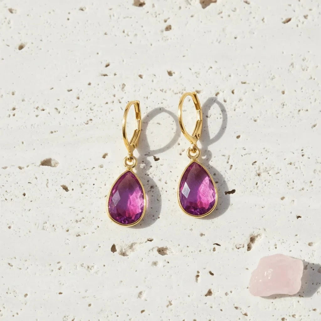 Gold earrings with pink teardrop-shaped gemstones on a textured beige surface.
