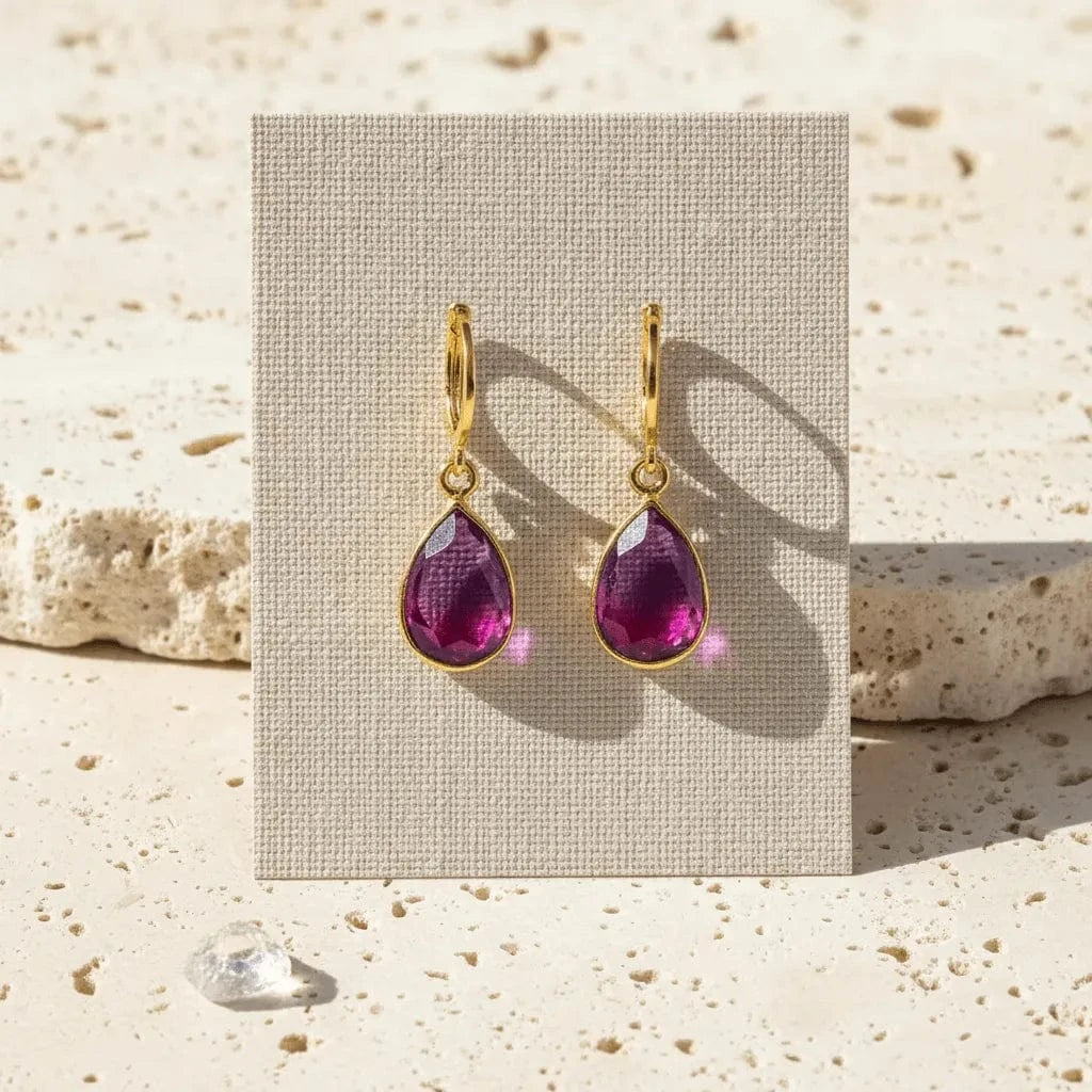 Gold earrings with purple teardrop stones on a beige card, placed on a textured stone surface.