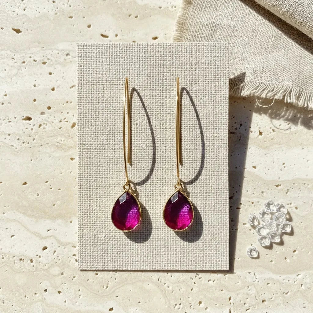 Gold earrings with pink teardrop stones on a beige card, with a textured beige surface in the background.