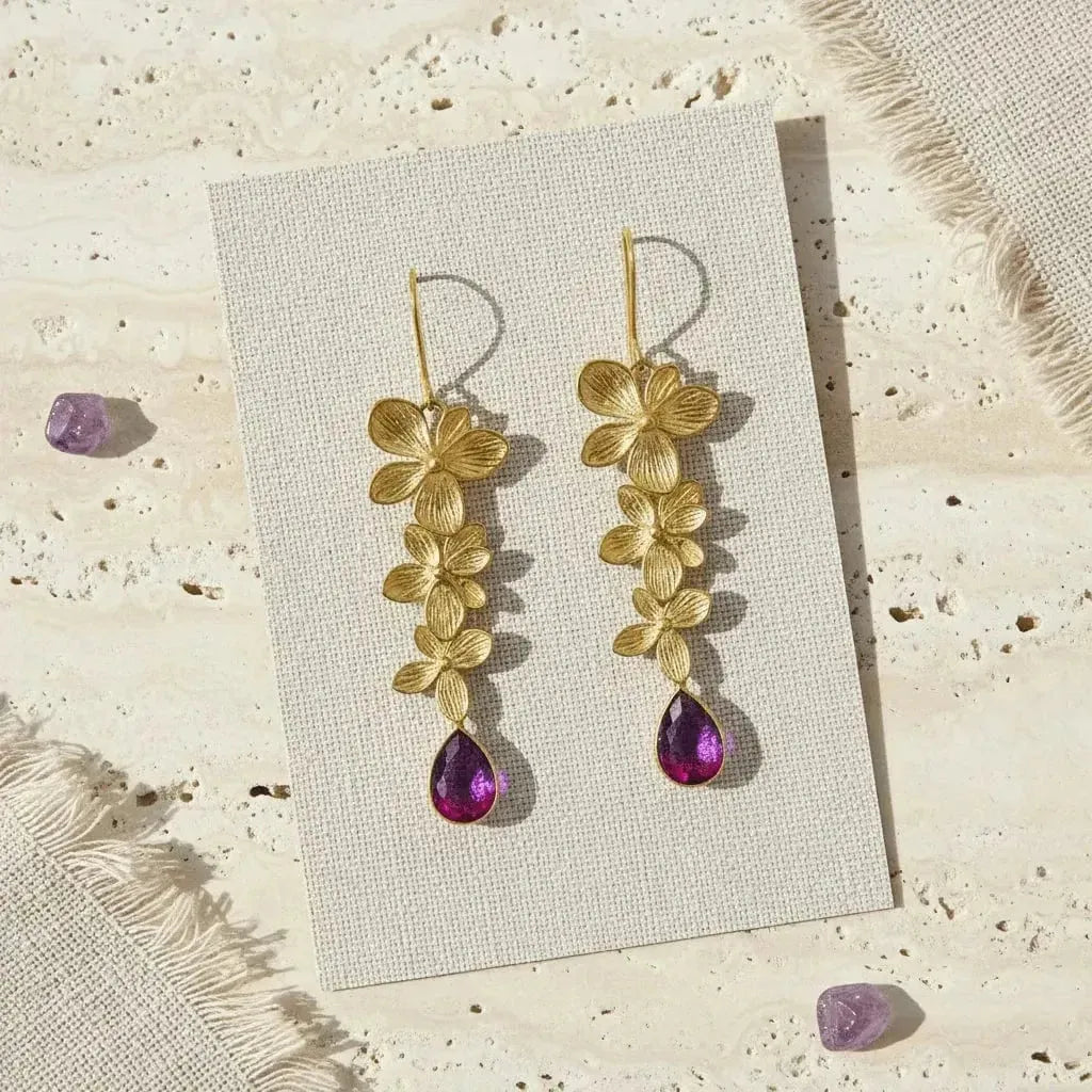 Gold floral earrings with purple teardrop stones on a textured card, surrounded by small purple stones.