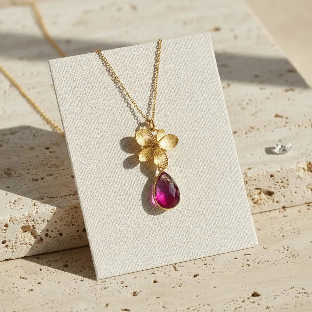 Gold necklace with a flower pendant and pink teardrop on a beige card.