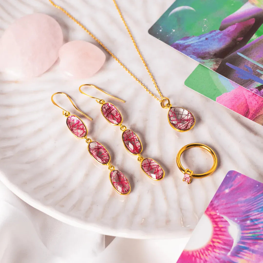 Gold jewelry set with pink gemstones on a shell with colorful cards in the background