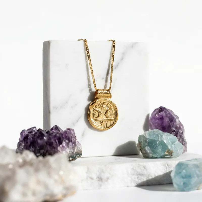 Gold necklace with a pendant on a white marble surface with amethyst and aquamarine crystals.