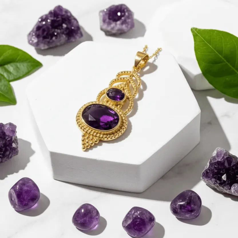Gold pendant with purple gemstones on a white stand, surrounded by amethyst crystals and green leaves.