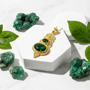 Gold pendant with green gemstones on a white stand surrounded by green leaves and crystals.