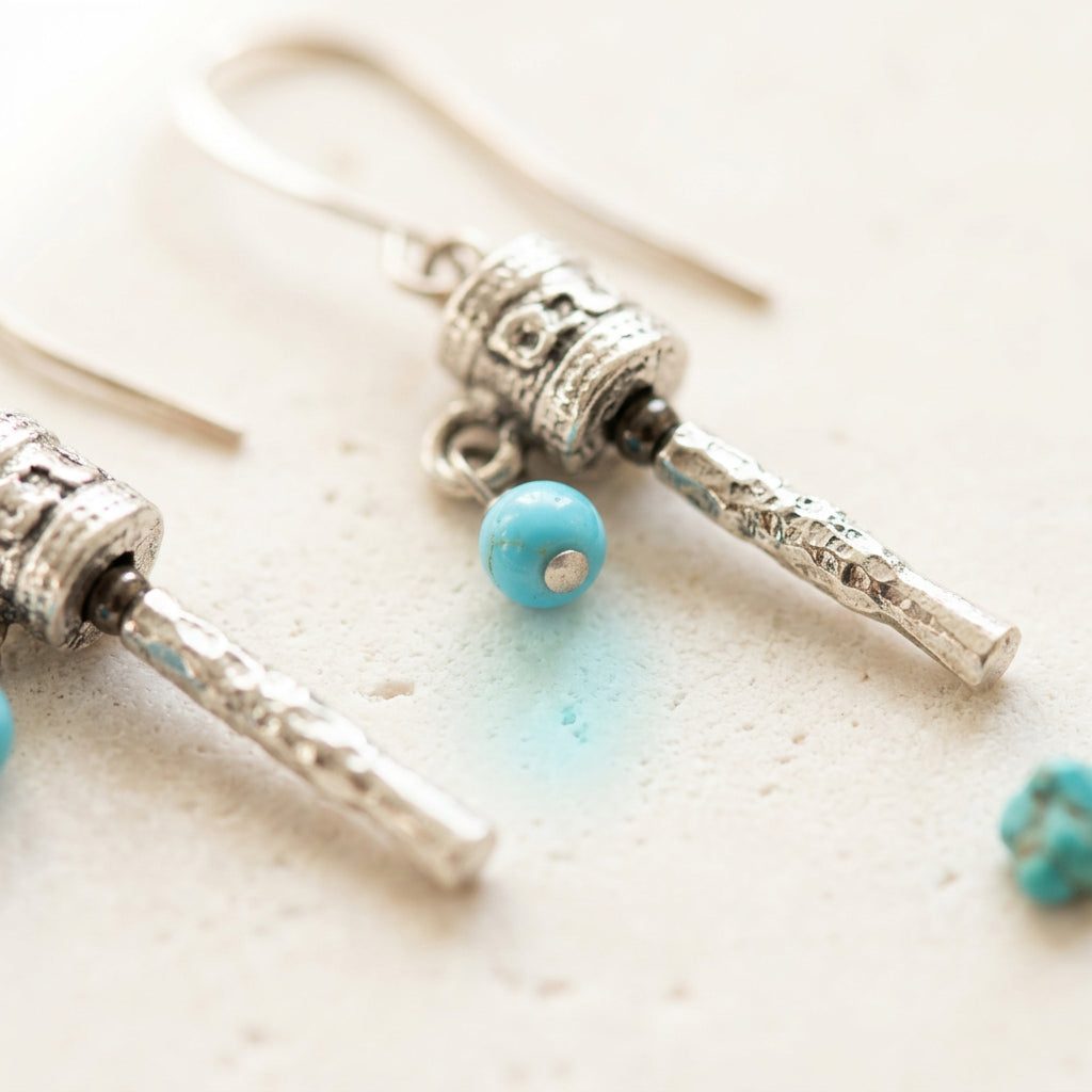 Close-up of a silver-plated prayer wheel earring showing the mantra drum and turquoise blue bead