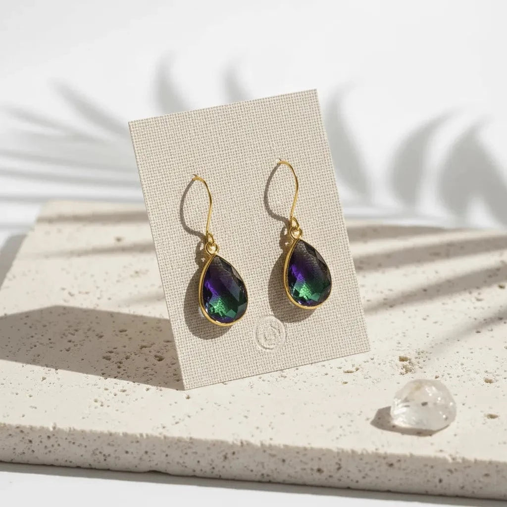 Teardrop-shaped earrings with gold setting on a beige card, placed on a textured surface.