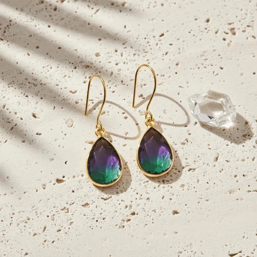 Gold earrings with purple and green teardrop-shaped gemstones on a textured beige surface.