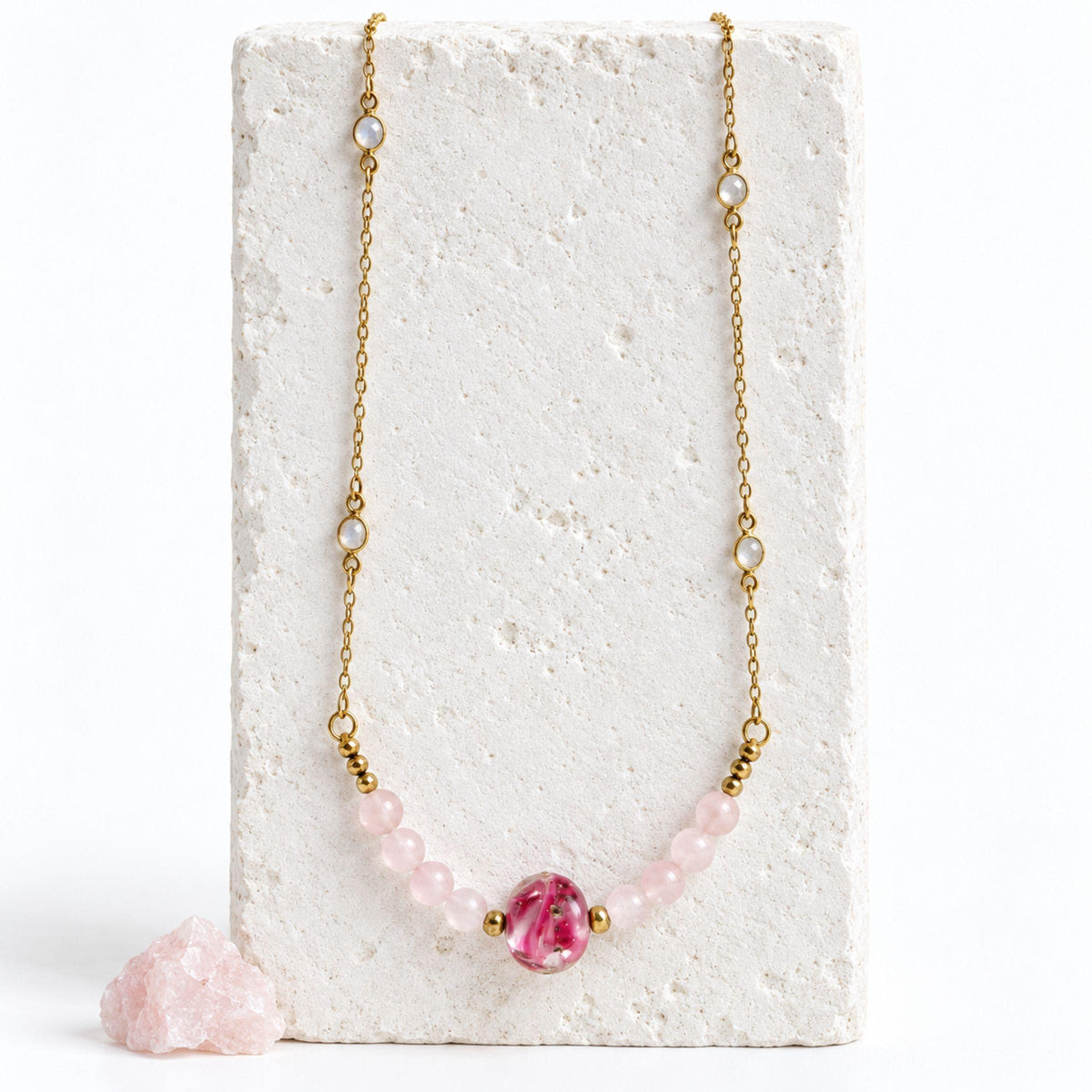 Rose quartz and moonstone necklace with a pink handblown glass focal and Ghana brass beads, draped on a white stone block beside raw rose quartz