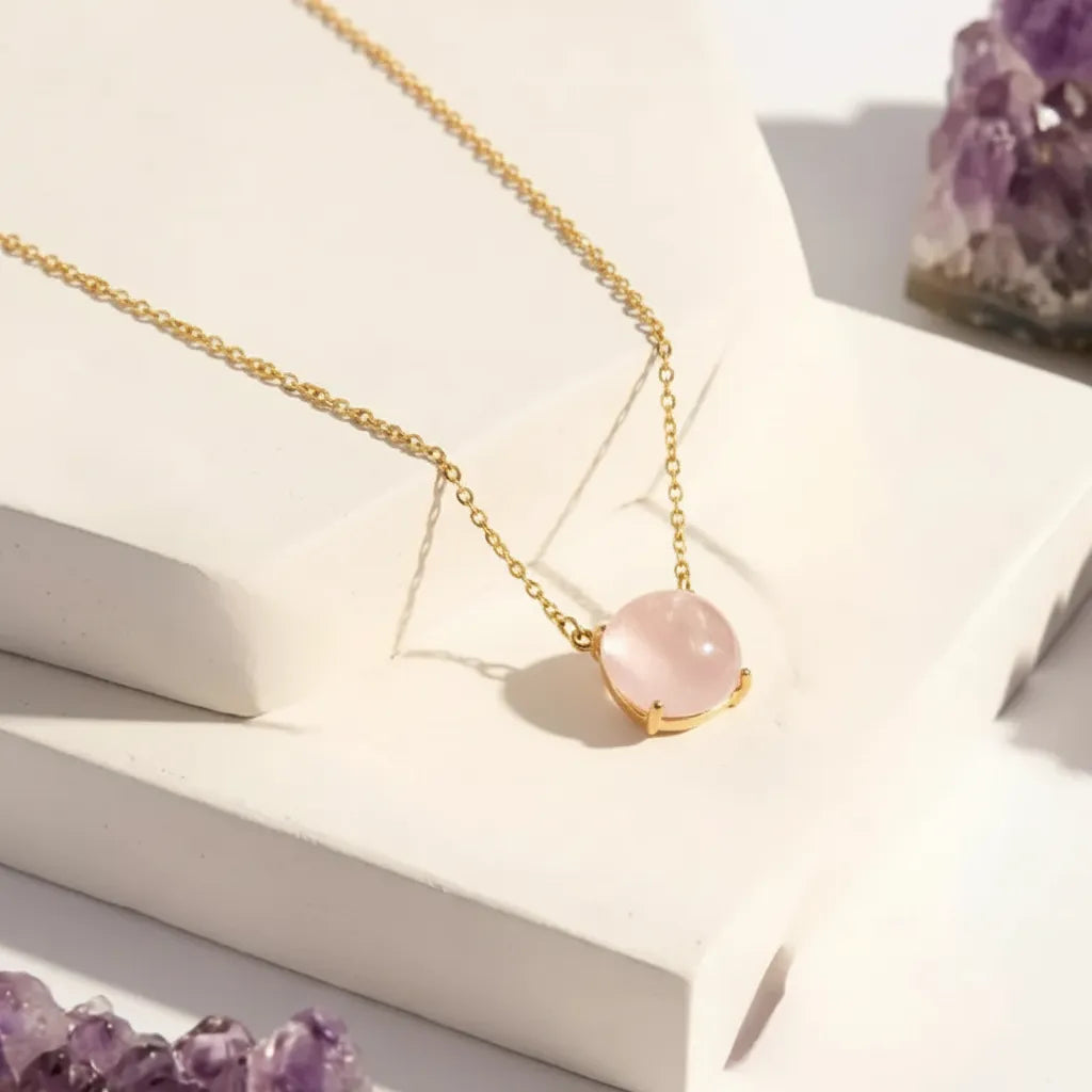 Gold necklace with a pink gemstone pendant on a white surface with purple crystals in the background.
