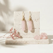 Pink teardrop earrings with gold hooks on a stone surface with pink flowers.