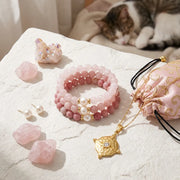Pink beaded bracelets, necklace, and crystals on a textured surface with a cat in the background.