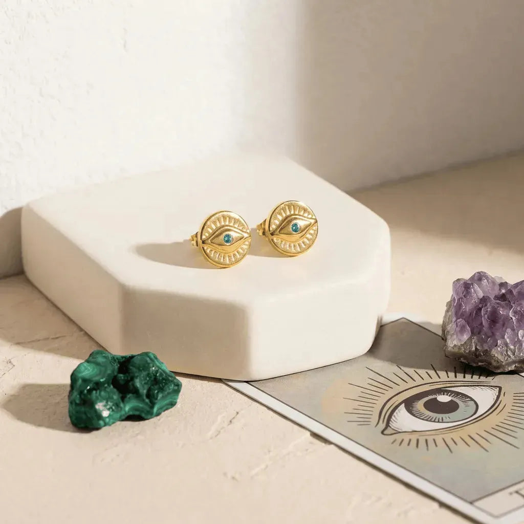 Gold earrings with eye design on a white stand with crystals and a card featuring an eye symbol.