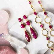 A pair of gold-plated statement earrings featuring three faceted rubies, displayed on a white surface with soft lighting.