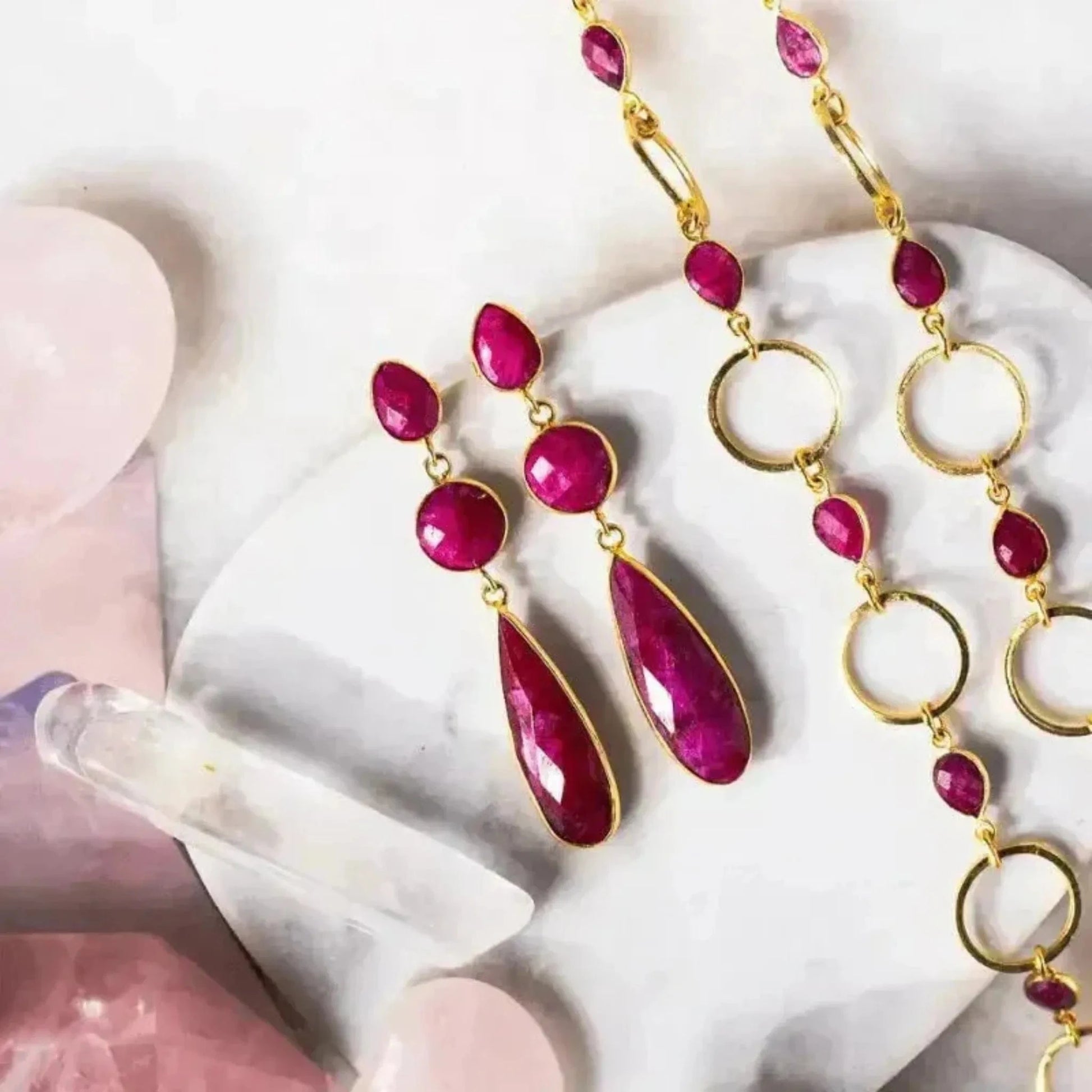 A pair of gold-plated statement earrings featuring three faceted rubies, displayed on a white surface with soft lighting.