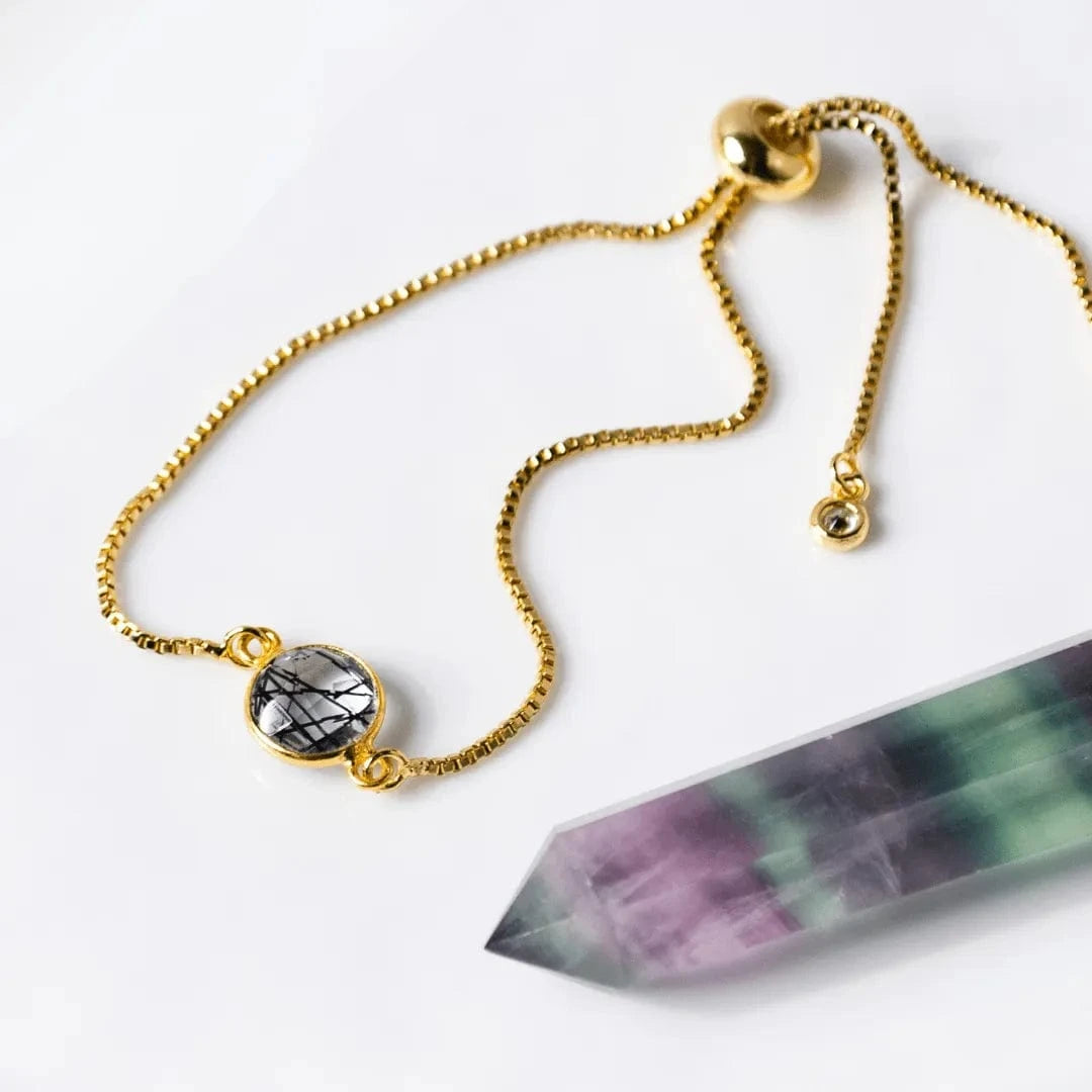 HUB Gold bracelet with a gemstone charm next to a crystal on a white background