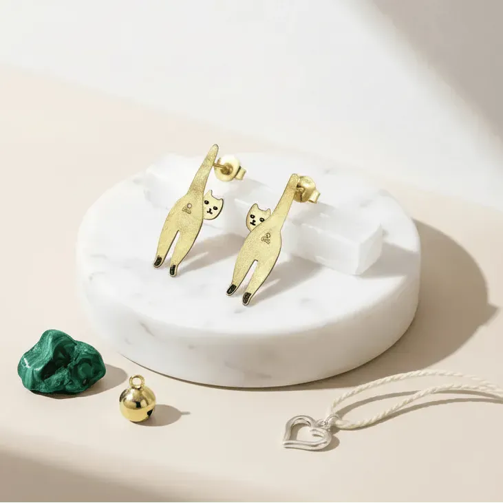 Gold cat-shaped earrings on a white dish with a green stone and silver necklace.