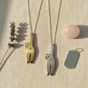 Two cat-shaped pendants on chains with a pink stone and gray tag on a beige surface.
