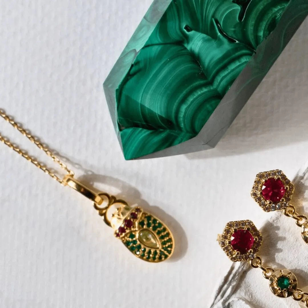 Gold jewelry with red and green gemstones on a white surface with a green marble-like object.