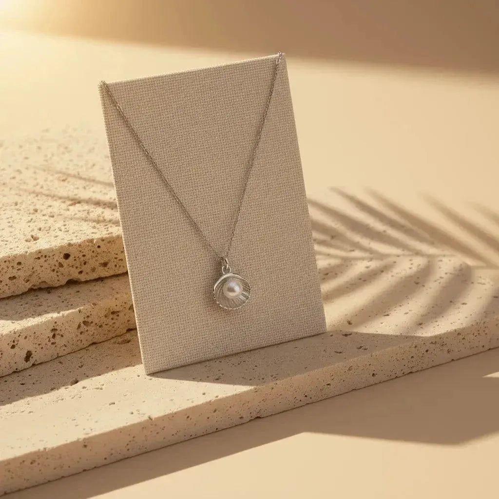 Necklace with a shell and pearl pendant on a sandy surface with a palm leaf shadow.