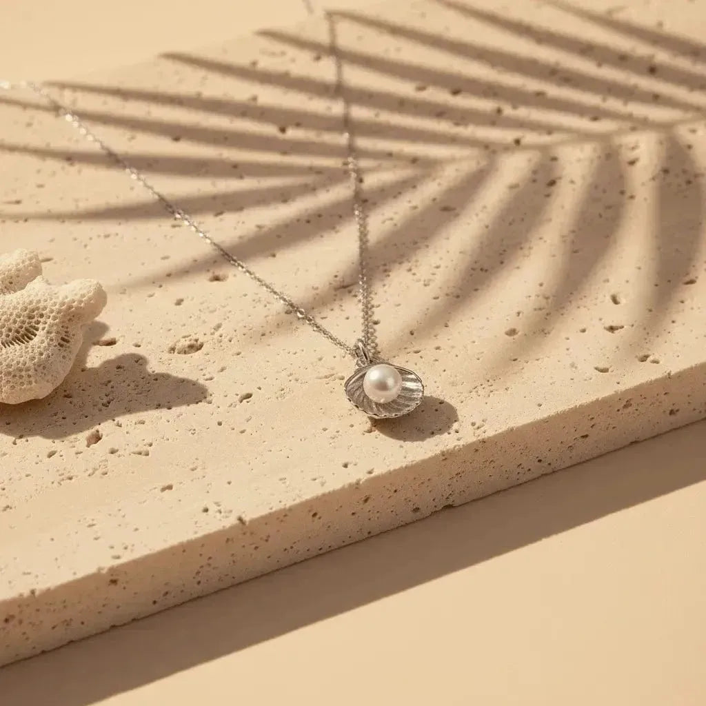 Necklace with a shell and pearl pendant on a sandy surface with a palm leaf shadow.